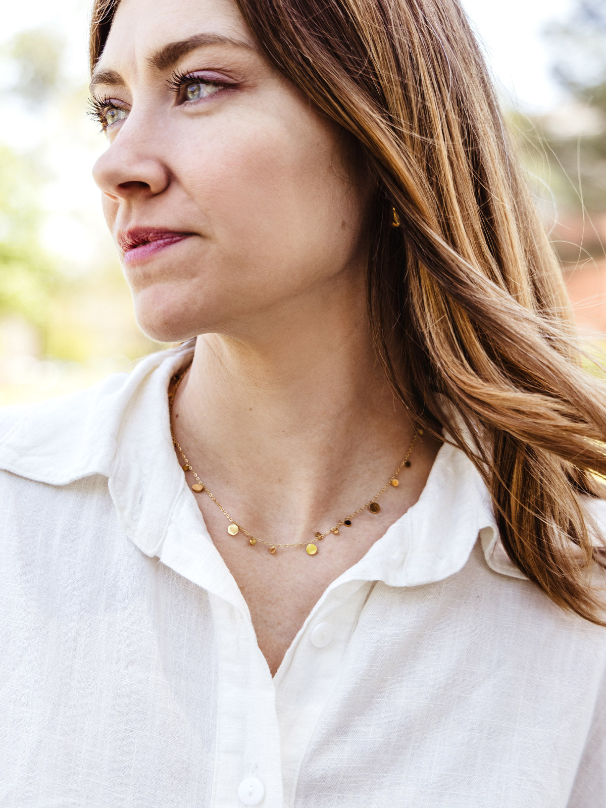 Close-up of a woman wearing a delicate gold disc necklace with a white collared shirt, looking off into the distance with natural lighting.