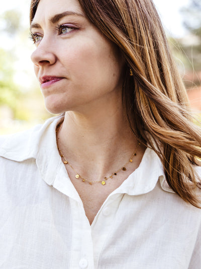 Close-up of a woman wearing a delicate gold disc necklace with a white collared shirt, looking off into the distance with natural lighting.