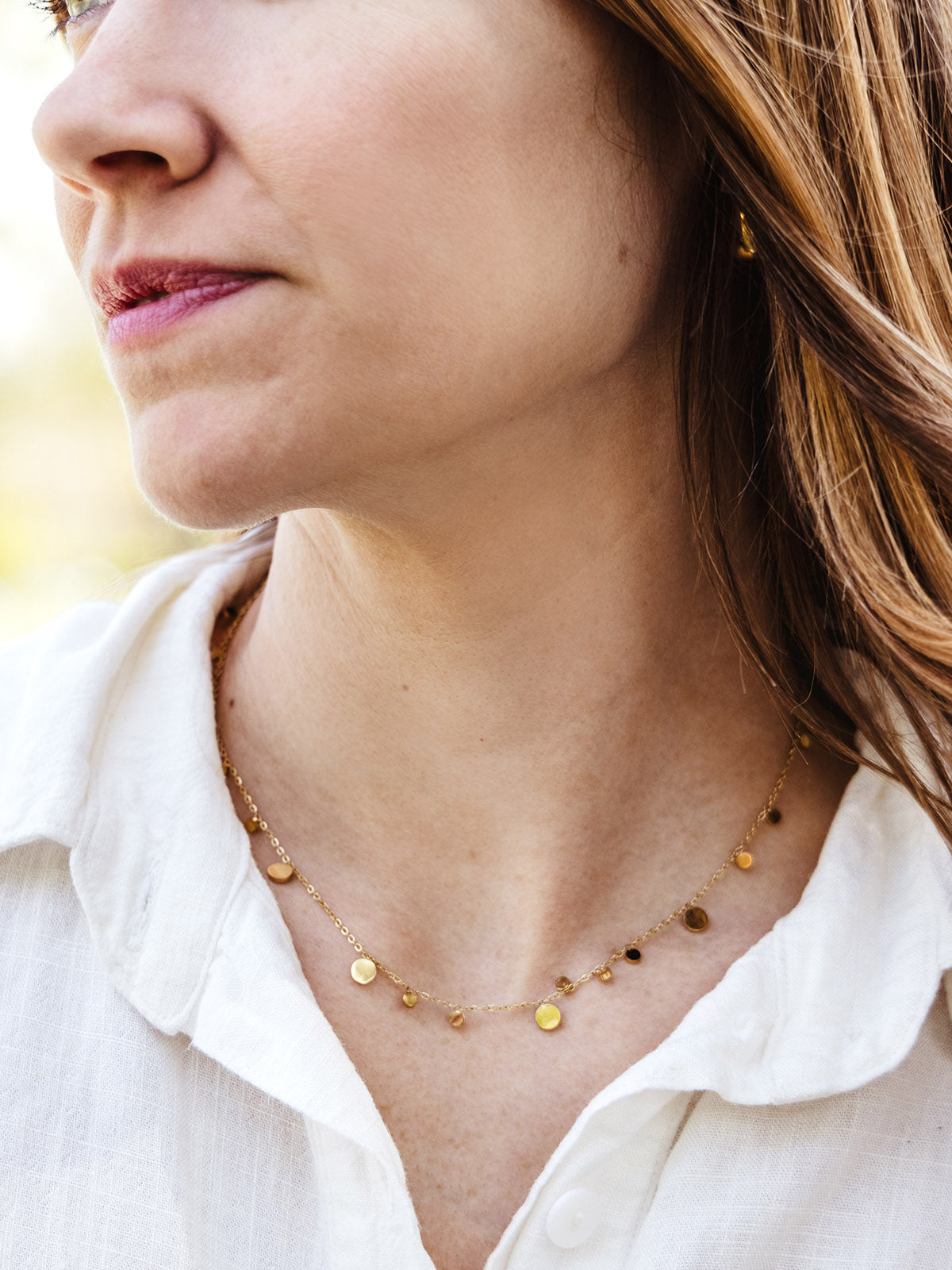 Close-up of a woman wearing a delicate gold necklace with small circular charms. She is dressed in a soft white button-up shirt, emphasizing minimalist and timeless artisan-made jewelry.