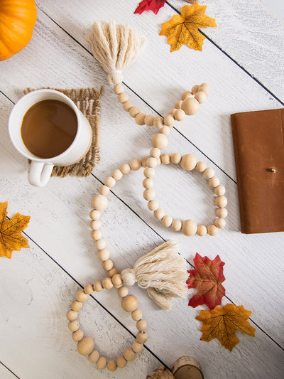 Neutral wooden bead garland with tassels arranged on a white wooden surface, styled with a cup of coffee on a woven coaster, scattered autumn leaves, a leather notebook, and a small pumpkin.