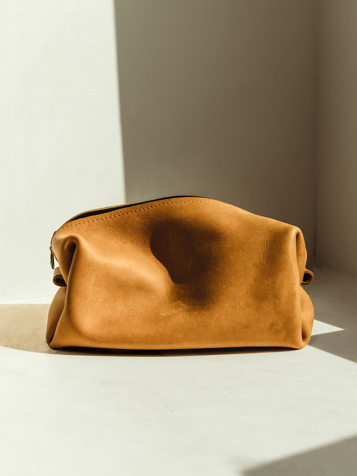  A soft, tan leather dopp kit sits in a sunlit corner on a white surface, casting a gentle shadow on the wall behind it.