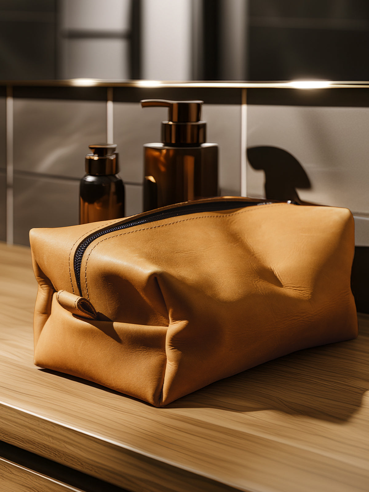 A structured tan leather dopp kit rests on a wooden bathroom counter with bronze pump bottles and tiled walls in the background.
