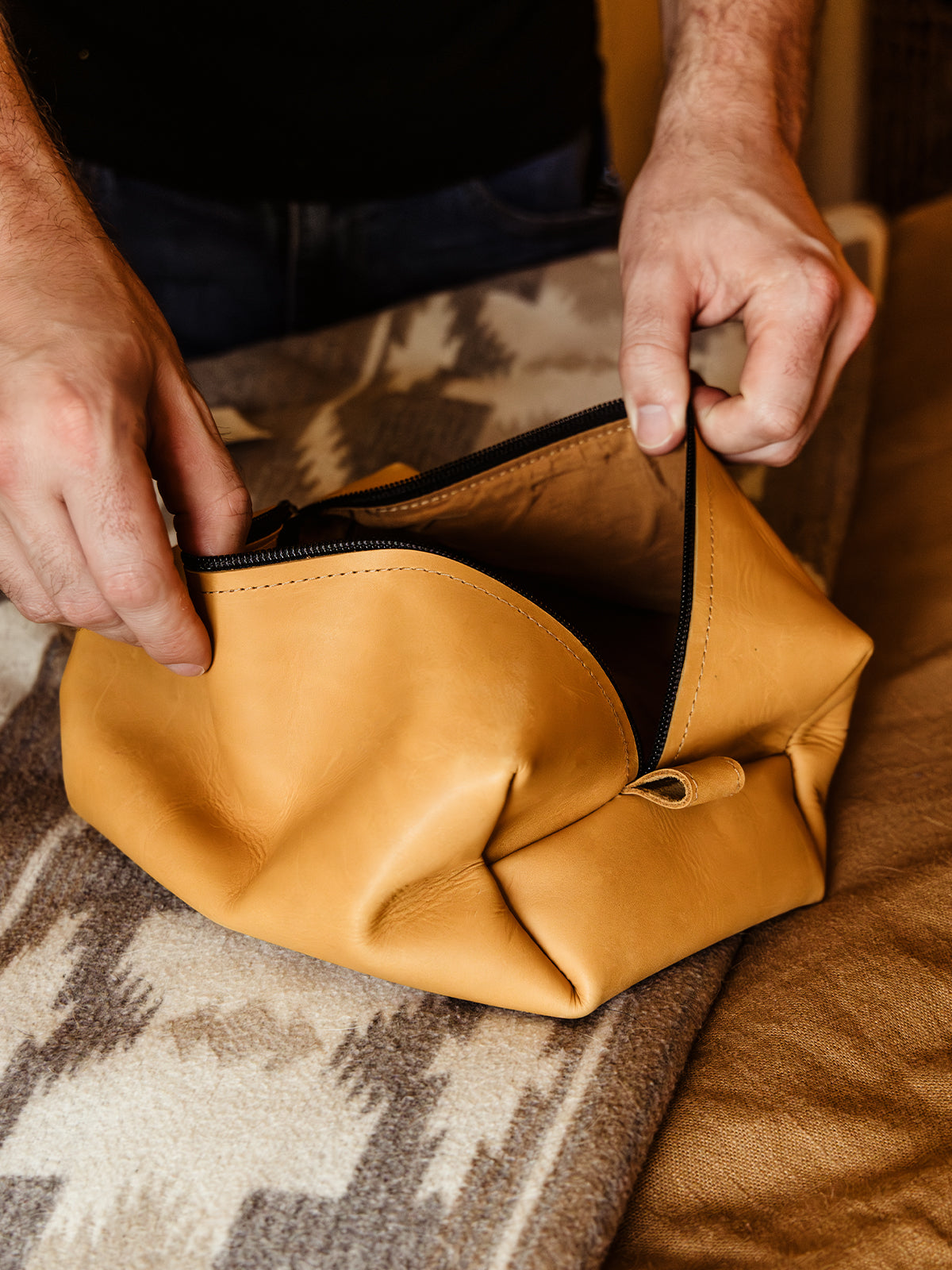 A person unzips a tan leather dopp kit on a patterned wool blanket, showing a close-up of their hands and the bag’s spacious interior.