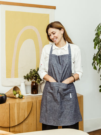 Smiling woman wearing a denim apron with front pockets, standing in a kitchen decorated with plants and artwork. Ethically made by Swahlee, perfect for cooking, available at Joffa.