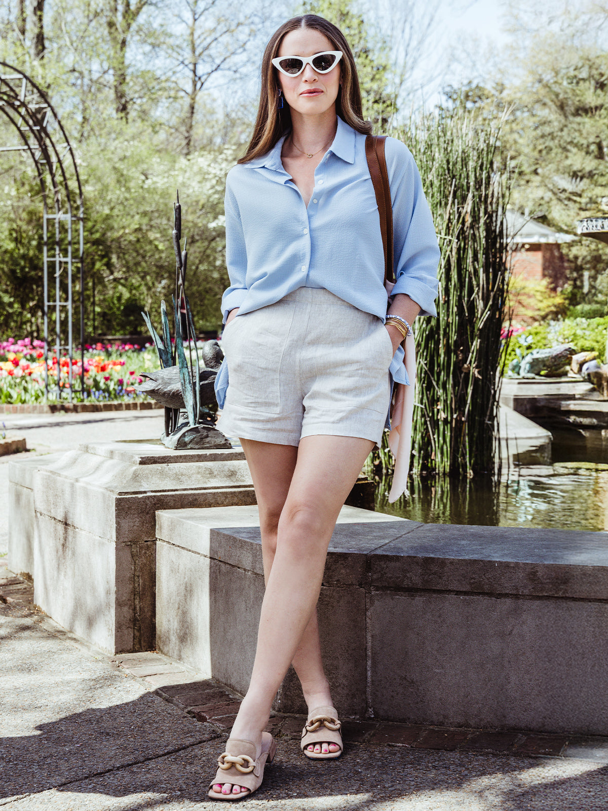 Full-body shot of a woman leaning against a low stone wall in a botanical garden. She wears a light blue blouse, natural linen shorts, nude slide heels, and white cat-eye sunglasses, accessorized with a brown leather tote and delicate jewelry.