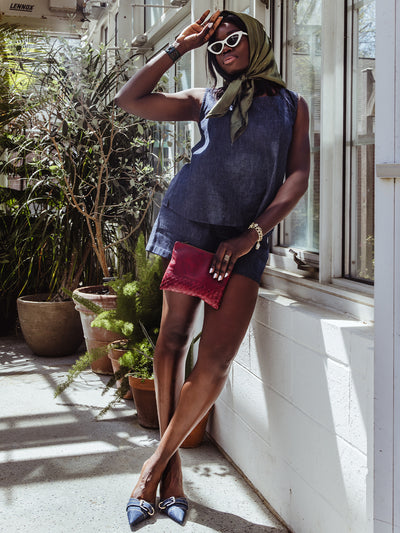 Full-body indoor shot of a woman in a denim sleeveless top and shorts. She pairs the look with pointed-toe denim heels, a green scarf tied around her head, and a red clutch bag.