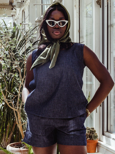 A woman wears a sleeveless denim set with a green silk scarf tied around her head and white cat-eye sunglasses. She stands indoors next to a sunlit window and potted plants, exuding retro elegance.