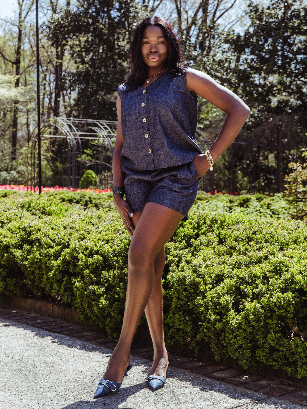 A woman poses outdoors in a sleeveless denim top with wooden buttons and matching high-waisted shorts. She wears pointed-toe denim heels and gold accessories, standing confidently in a garden path.