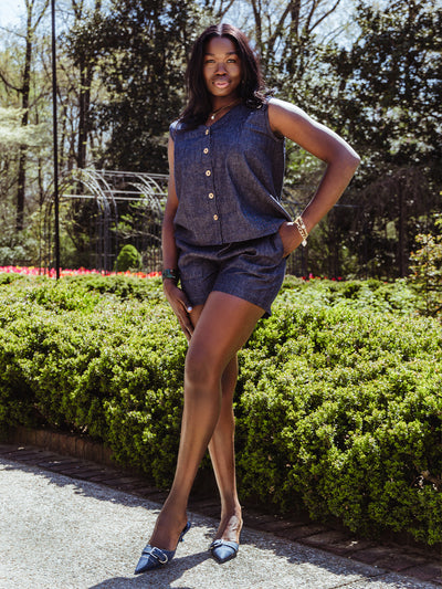 A woman poses outdoors in a sleeveless denim top with wooden buttons and matching high-waisted shorts. She wears pointed-toe denim heels and gold accessories, standing confidently in a garden path.
