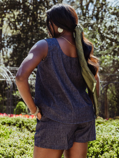 Rear view of a woman wearing a dark denim tank and shorts set, styled with a long green hair scarf, gold earrings, and layered bracelets. She stands in front of blooming garden greenery.