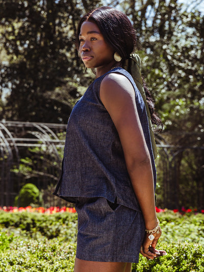Side profile of a woman modeling a sleeveless denim top and shorts, with subtle side vents on the top and minimalist accessories. Her hair is styled with a sage green scarf.
