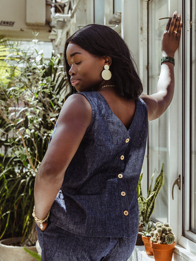 Close-up from behind of a woman in a sleeveless denim top featuring a button-down back. She accessorizes with large gold statement earrings, a chain bracelet, and a gold necklace.