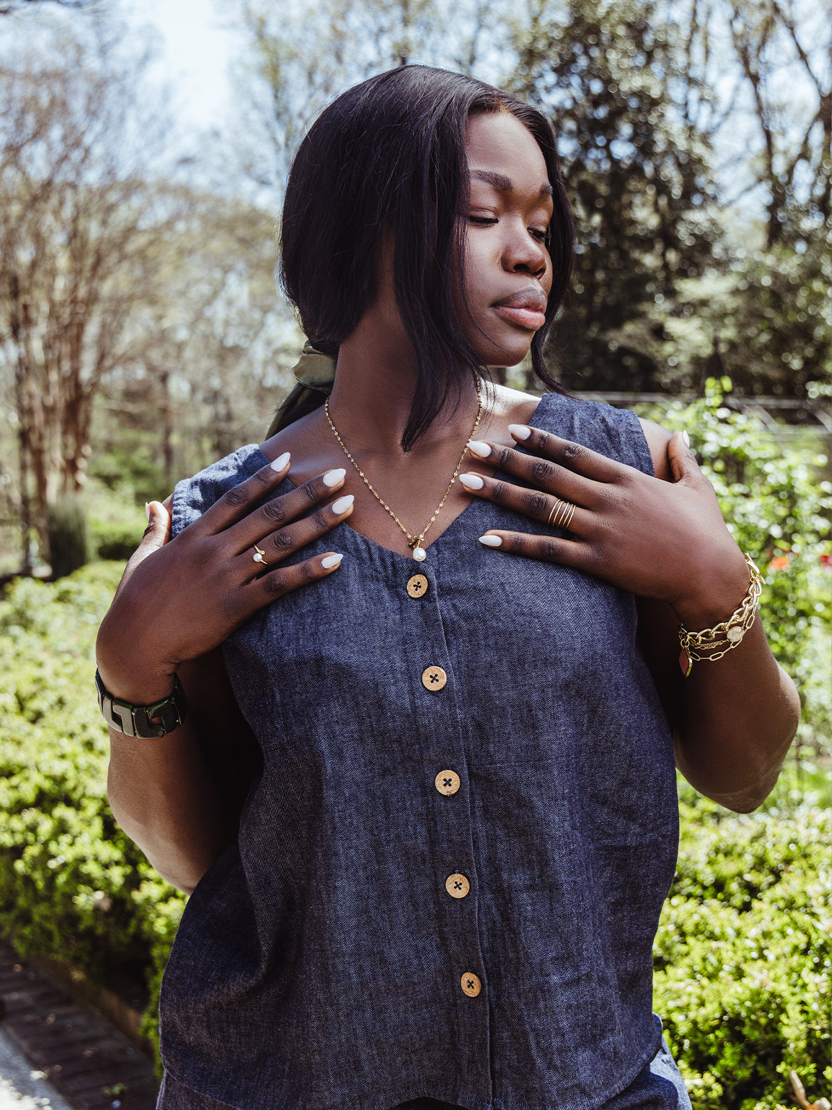 Close-up of a woman outdoors wearing a button-front denim top. She holds her hands near her collar, showing off gold rings, a pearl charm necklace, a chunky bracelet, and soft neutral nails.