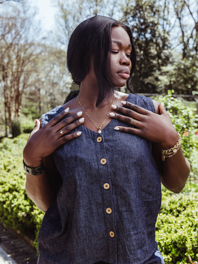 Close-up of a woman outdoors wearing a button-front denim top. She holds her hands near her collar, showing off gold rings, a pearl charm necklace, a chunky bracelet, and soft neutral nails.
