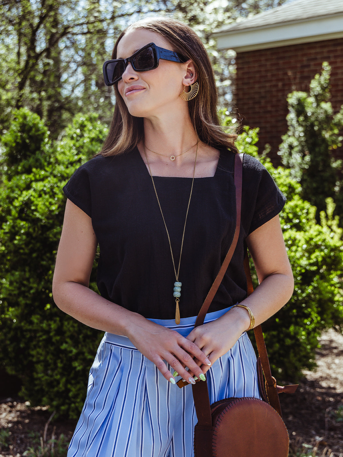 lose-up of a woman in a black square-neck top and striped pants, accessorized with a brown crossbody bag, bold gold earrings, and layered jewelry. She wears oversized black sunglasses and stands in front of a hedge-lined path.