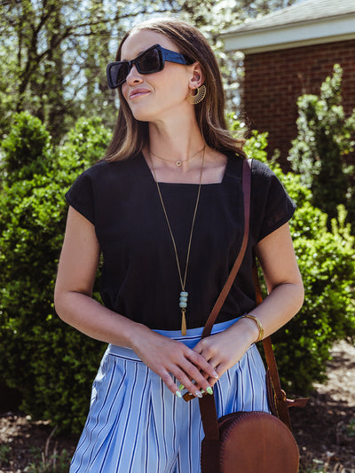 lose-up of a woman in a black square-neck top and striped pants, accessorized with a brown crossbody bag, bold gold earrings, and layered jewelry. She wears oversized black sunglasses and stands in front of a hedge-lined path.