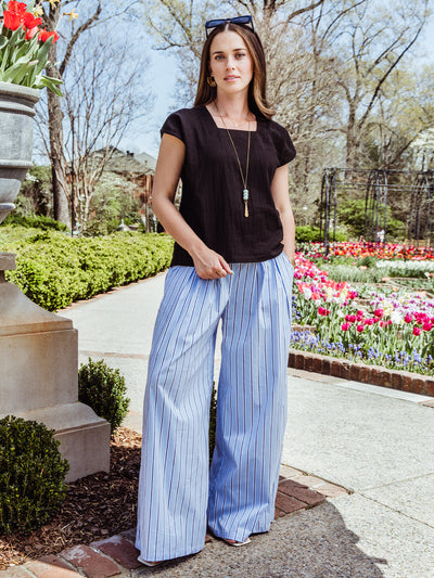 Full-body image of a woman in a black square-neck blouse and flowing blue striped wide-leg pants. She stands in a garden pathway lined with tulips, styled with sunglasses and layered gold jewelry.