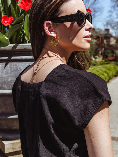 Close-up of a woman wearing a black square-neck top, gold fan-shaped earrings, and black oversized sunglasses. Her back is turned slightly to the camera, revealing a gold chain necklace and bright red tulips in the background.