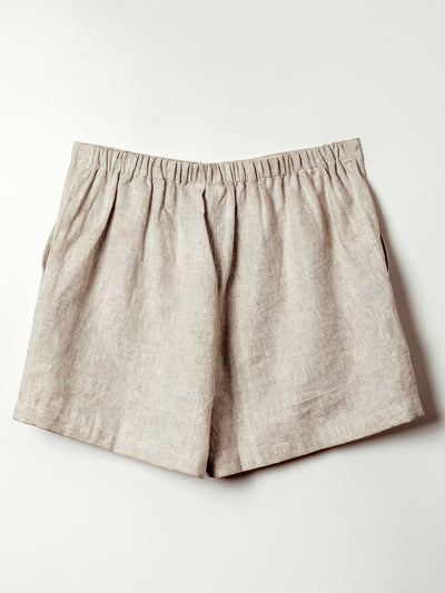A pair of beige linen shorts is laid flat against a light background. These shorts feature an elastic waistband and a softly gathered silhouette, creating a relaxed and breathable fit ideal for warm weather.