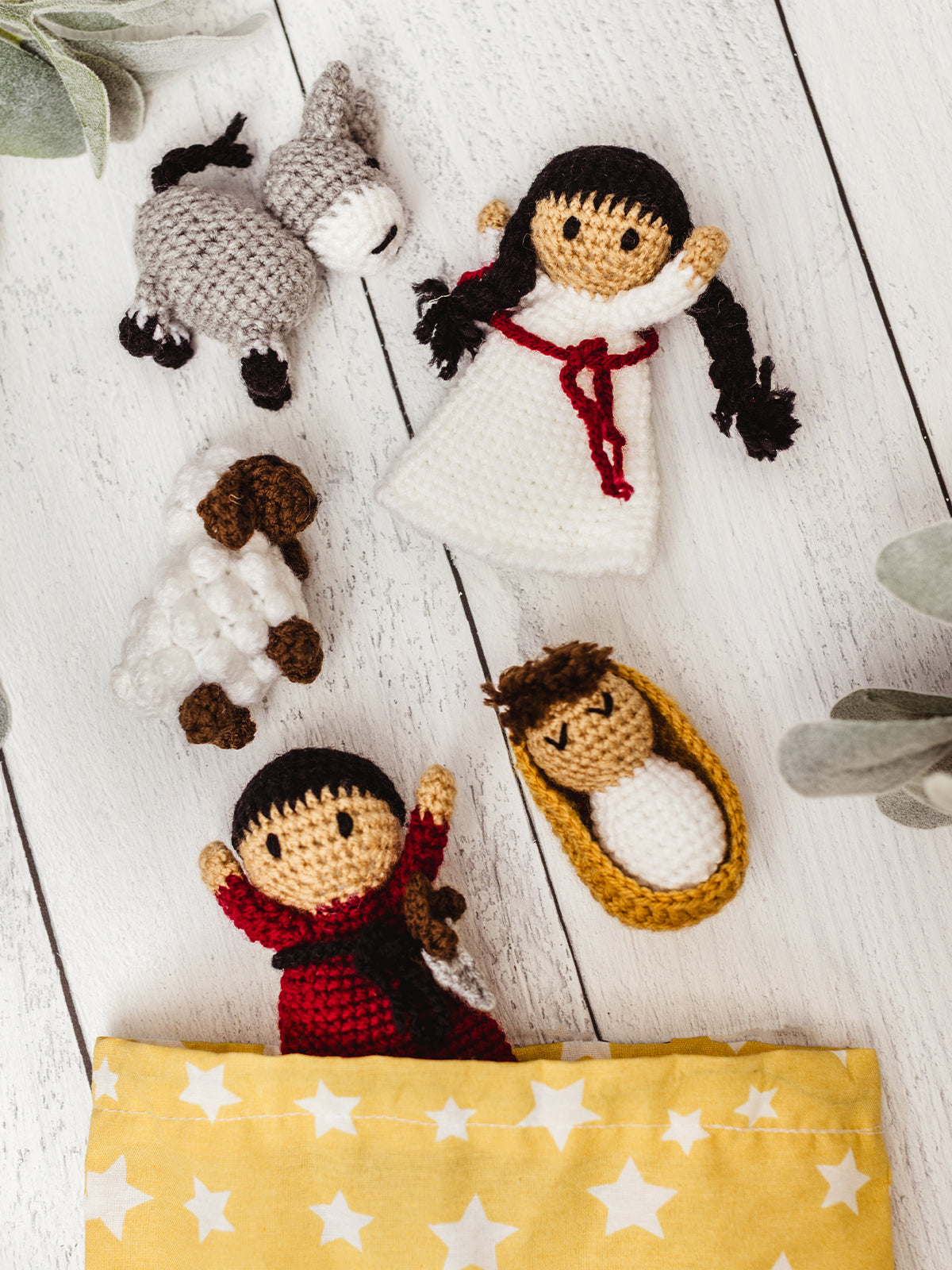 Flat lay of handmade crochet Nativity set including Mary, Joseph, baby Jesus in a manger, a sheep, and a donkey on a white wood background. Fair trade Christmas decor and play set.