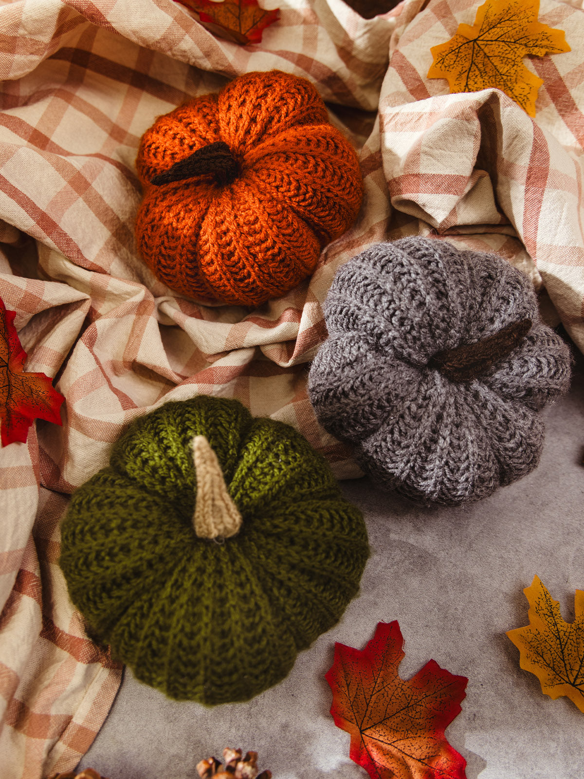 Three knitted pumpkins in fall colors—orange, green, and gray—arranged on a plaid blanket with autumn leaves, creating a cozy seasonal decor scene.
