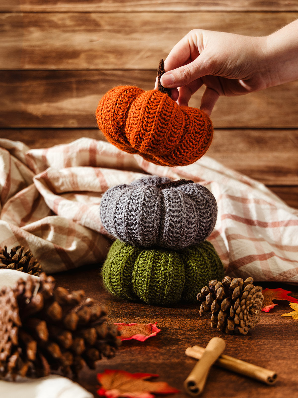 Hand stacking knitted pumpkins in orange, gray, and green on a wooden table with pinecones, cinnamon sticks, and fall leaves."