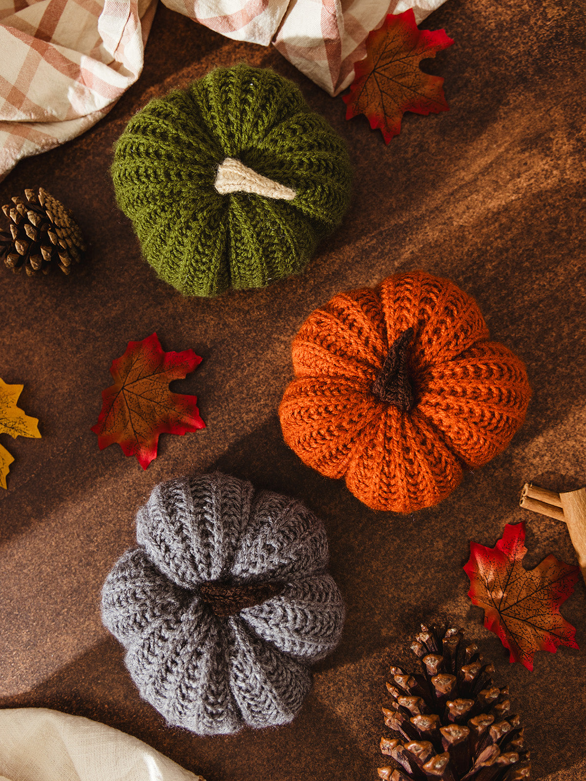 Flat lay of three knitted pumpkins in green, orange, and gray surrounded by pinecones, cinnamon sticks, and autumn leaves on a rustic wooden surface.