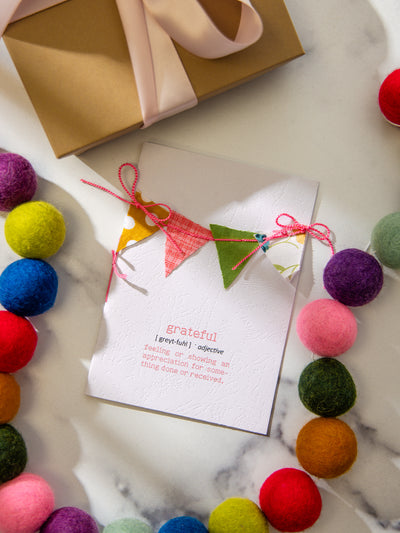 Handmade “Grateful” definition card with colorful fabric bunting and pink string bows, styled flat on marble with a kraft gift box and felt ball garland in bright rainbow colors.