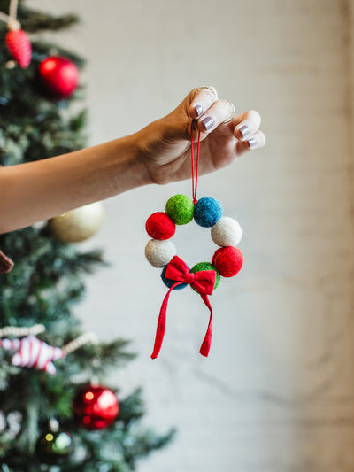 Hand holding a colorful felt ball Christmas wreath ornament with a red ribbon bow, ready to hang on a holiday tree — handmade festive décor.
