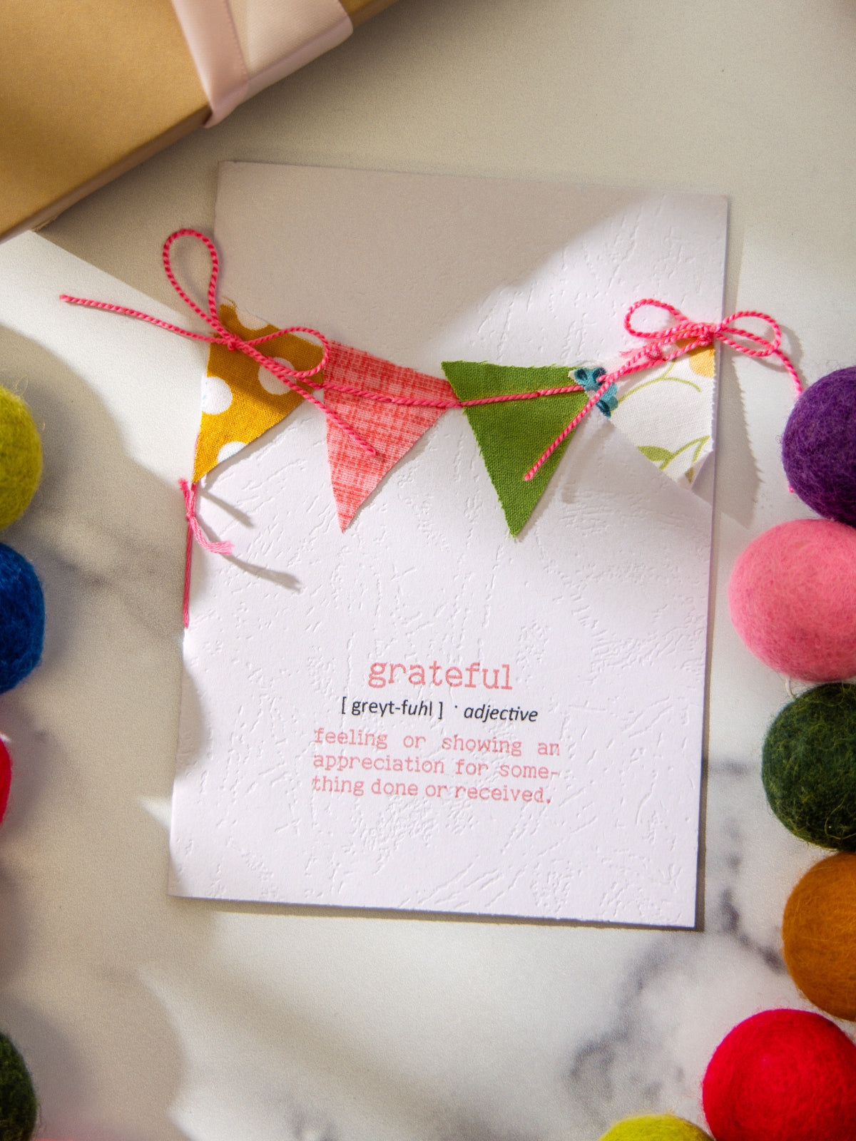Close-up of white textured card featuring the word grateful in red with dictionary-style definition, decorated with fabric bunting and pink bows, surrounded by felt pom garland and a wrapped gift.