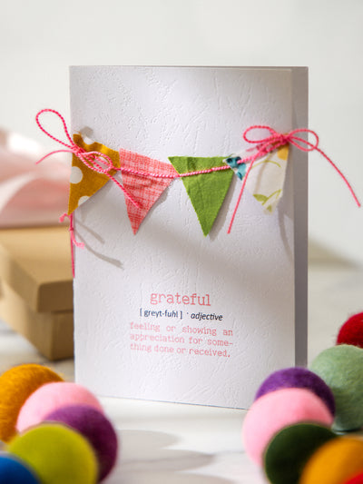 Standing view of handmade “Grateful” definition card with colorful bunting banner across the top, styled with felt pom pom garland and a wrapped kraft gift in the background.