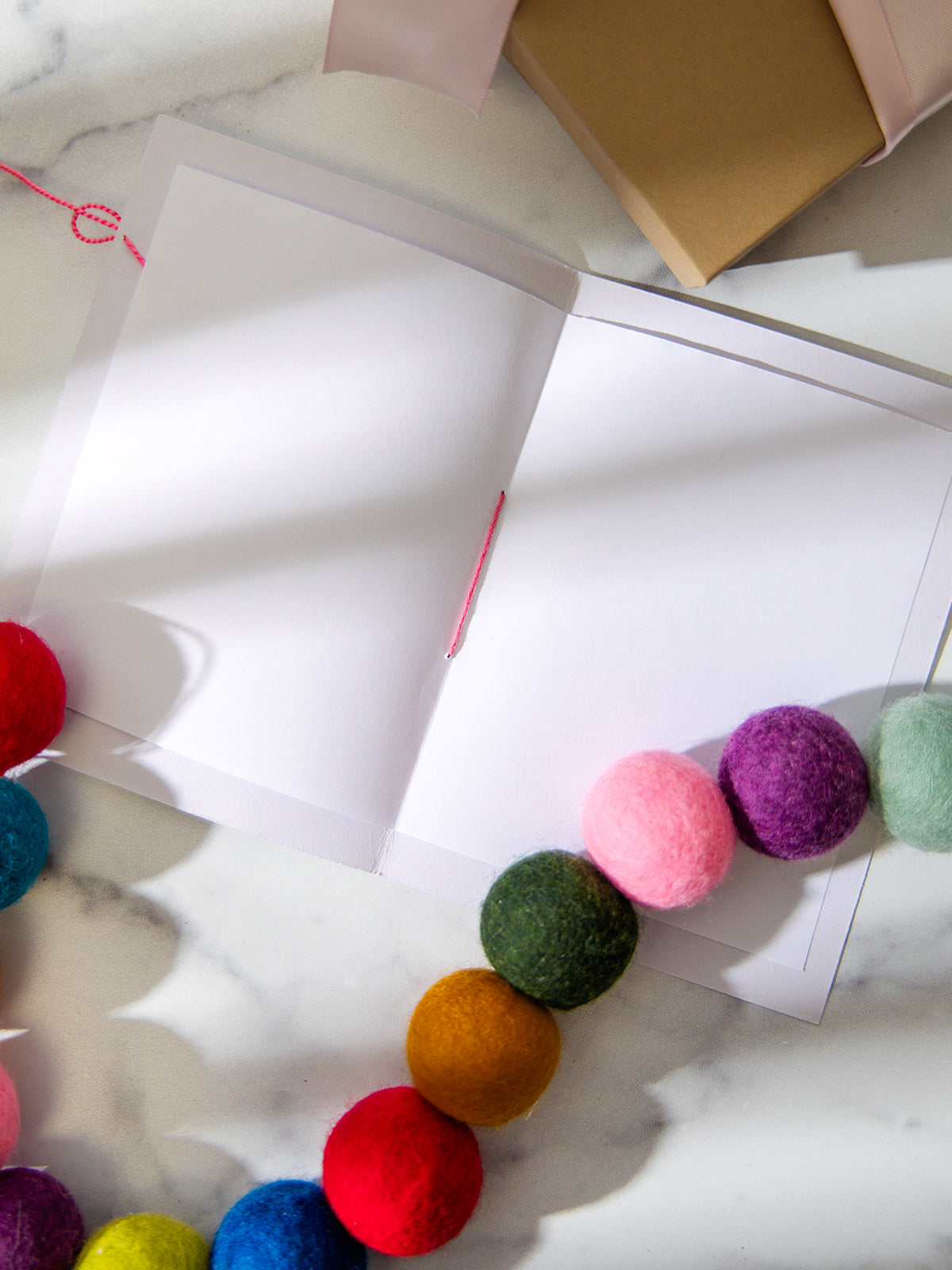 Inside of blank “Grateful” handmade card displayed on marble surface, styled with rainbow felt pom pom garland and kraft gift box in the corner.