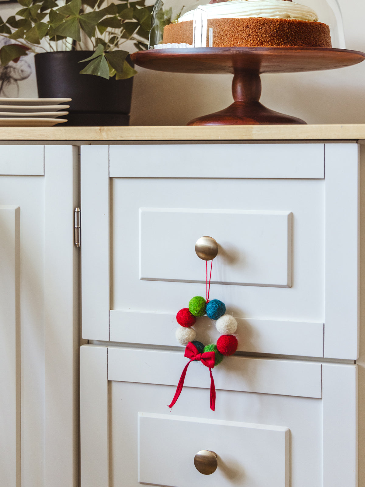 Colorful felt ball Christmas wreath ornament with a red ribbon bow hanging on a white kitchen cabinet knob beside a cake stand — handcrafted holiday home décor.