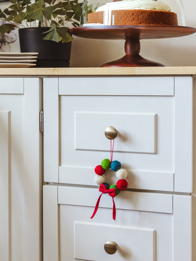 Colorful felt ball Christmas wreath ornament with a red ribbon bow hanging on a white kitchen cabinet knob beside a cake stand — handcrafted holiday home décor.
