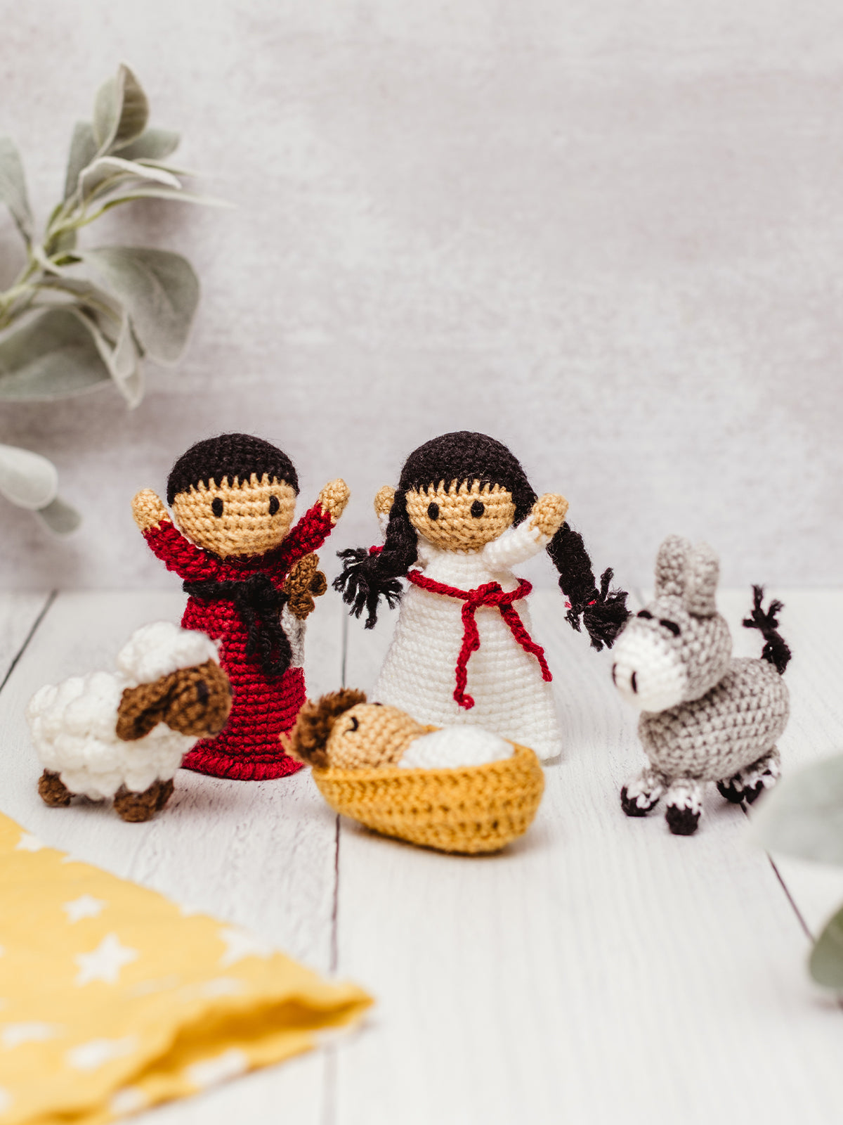 Handcrafted crochet Nativity scene with Mary, Joseph, baby Jesus in a manger, donkey, and sheep arranged on a white wooden surface. Fair trade Christmas decoration and kids’ play set.