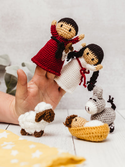 Hand holding handmade crochet Nativity finger puppets with Mary and Joseph, alongside a crocheted baby Jesus in a manger, sheep, and donkey. Ethical fair trade Christmas toy set for kids and holiday storytelling.