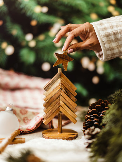 Person decorating a rustic wooden Christmas tree ornament by adding a star topper, sustainable handmade holiday décor surrounded by pinecones and cozy textiles.