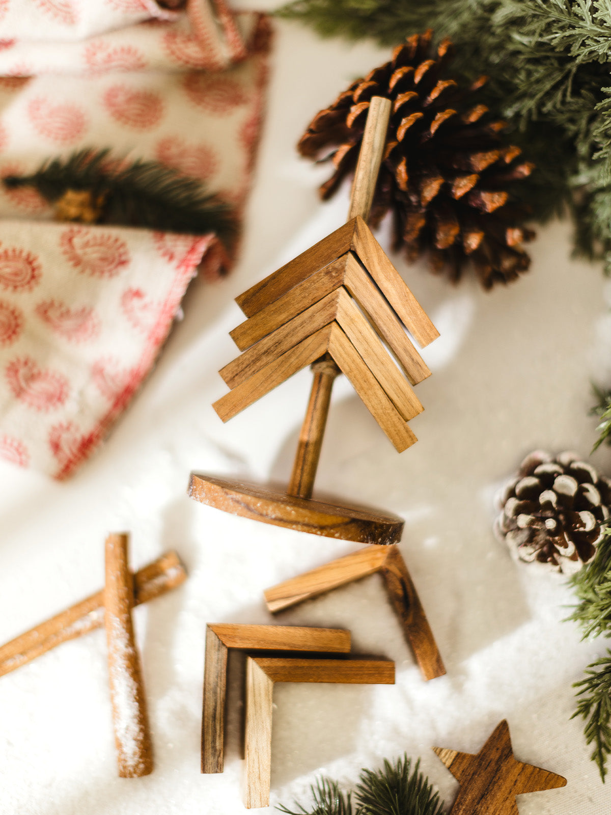 Flat lay of a wooden Christmas tree decoration being assembled with detachable wood pieces, star topper, and natural pinecones on a snowy holiday background.