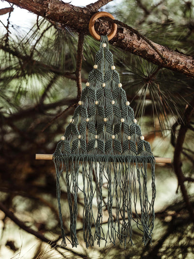 Handmade macramé Christmas tree wall hanging in green, adorned with wooden beads and tassels, suspended from a wooden ring on a pine tree branch.