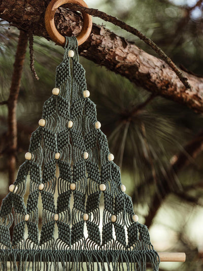 Close-up view of a green macramé Christmas tree wall hanging with intricate knots and wooden beads, hanging from a wooden ring on a natural pine branch.