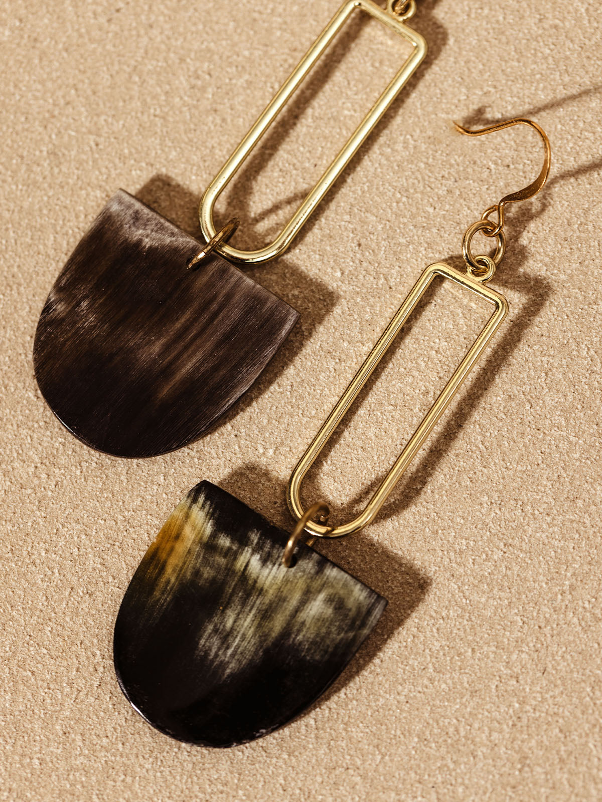 Pair of long drop earrings with rectangular gold frames and dark horn pendants, laid flat on a light tan surface, showcasing their unique geometric design and natural material.