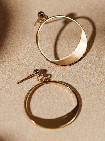 Pair of gold crescent-shaped hoop earrings laid flat on a light tan surface, highlighting their minimalist design and smooth finish.