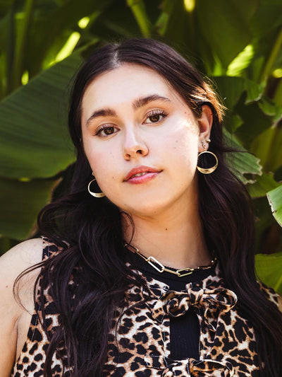 Close-up of a woman wearing gold crescent-shaped hoop earrings, paired with a leopard print top and a gold chain necklace, standing against a backdrop of green leaves.