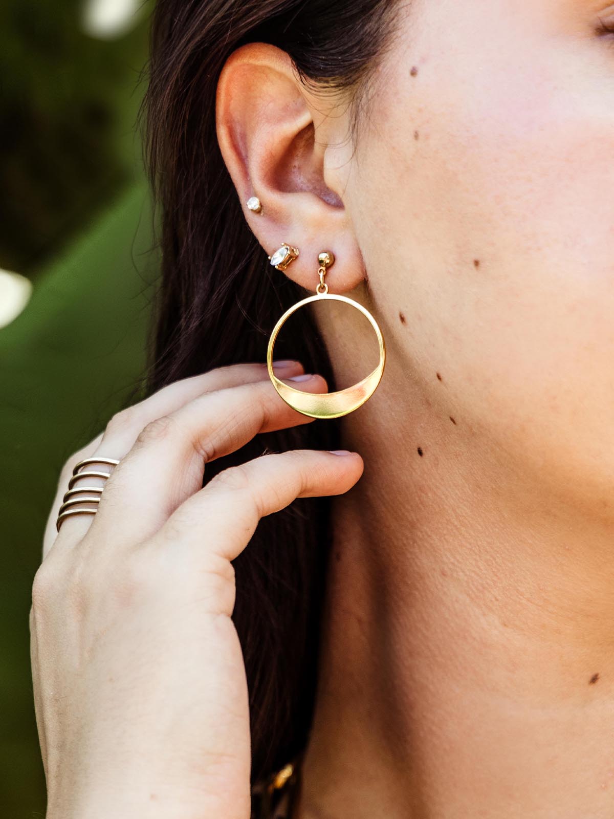 Close-up of a woman's ear wearing a gold crescent-shaped hoop earring, with her hand gently touching the earring, showcasing her gold ring and multiple ear piercings.