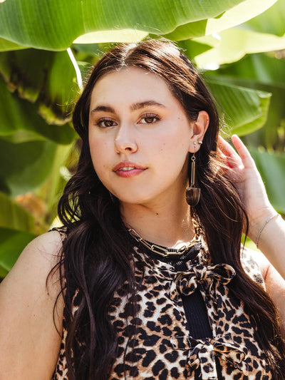 Woman wearing long, rectangular drop earrings with a dark horn pendant at the bottom, paired with a leopard print top and standing against a backdrop of large green leaves.