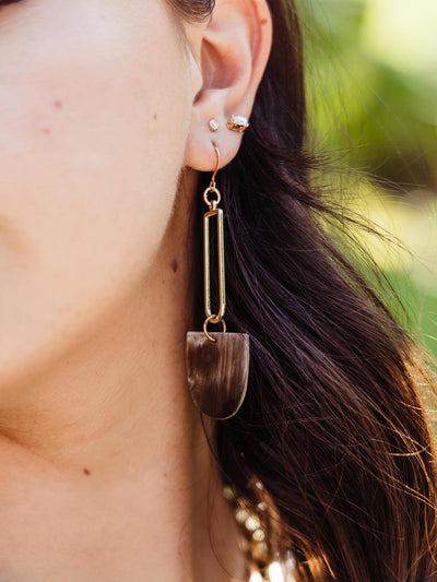 Close-up of a woman's ear adorned with a long, rectangular drop earring featuring a dark horn pendant, highlighting her multiple ear piercings and soft brown hair.