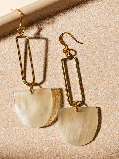 Close-up of artisanal drop earrings with gold rectangular frames and natural horn pendants, styled against a neutral backdrop.