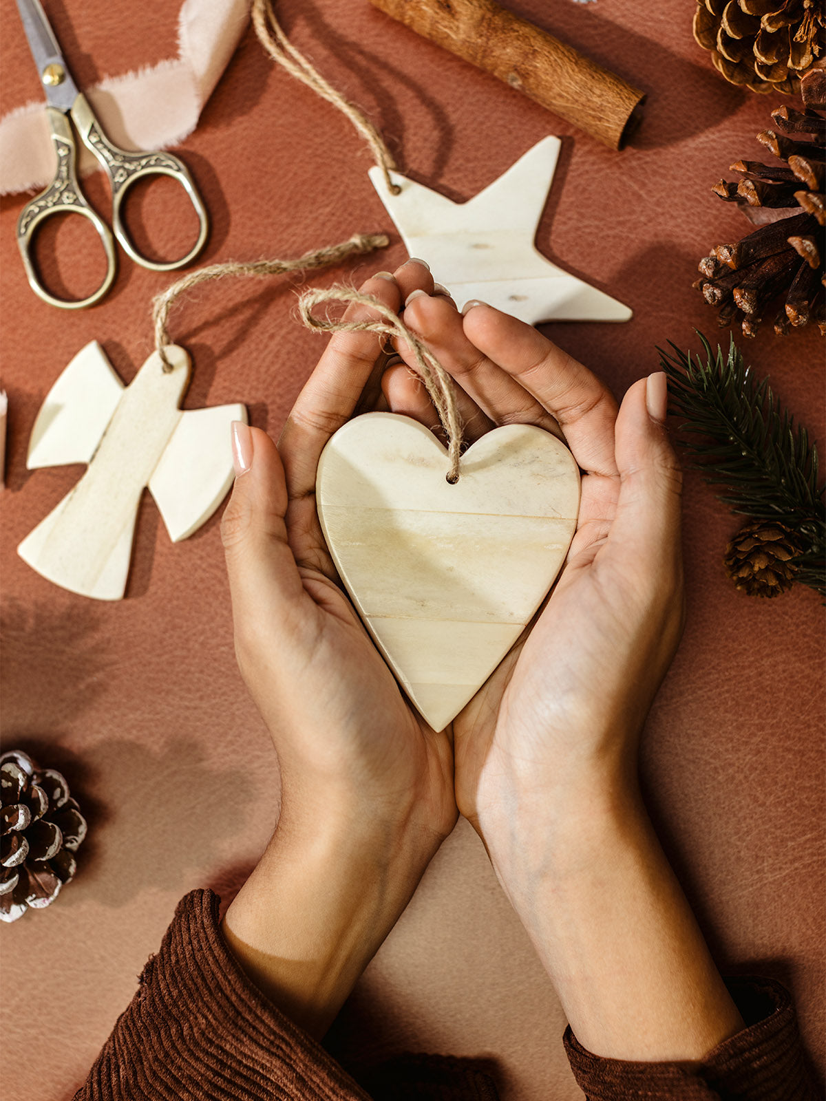 Hands holding a handmade bone heart ornament with jute string, sustainable Christmas décor surrounded by bone star and angel ornaments, pinecones, and festive accents.