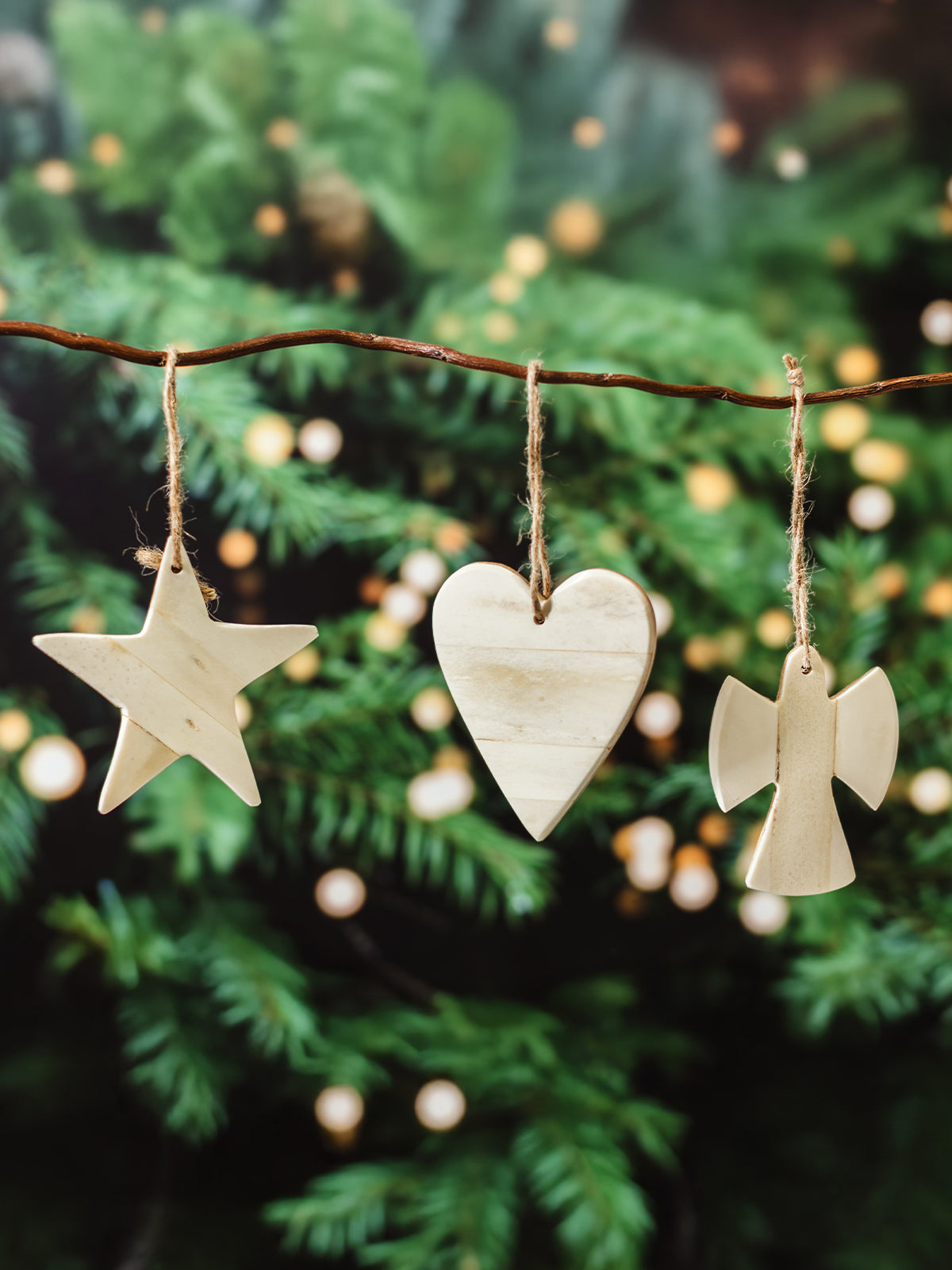 Set of three natural bone Christmas ornaments — star, heart, and angel — hanging on twine from a rustic branch in front of a decorated holiday tree.