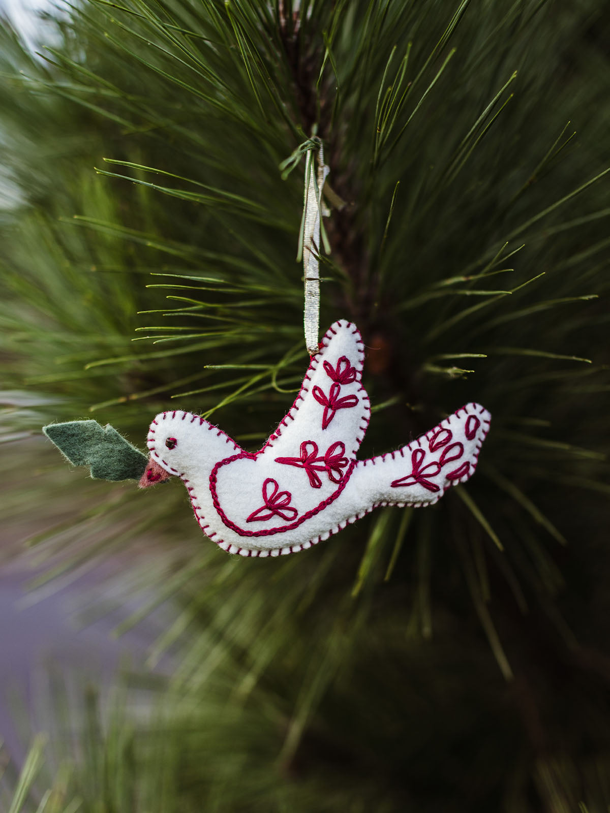 Handmade white felt dove ornament with red embroidery and green leaf detail hanging on Christmas tree branch – eco-friendly holiday decor